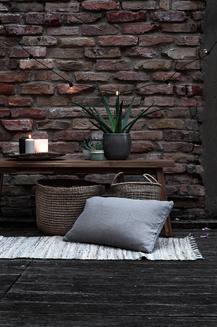 Holzbank mit Pflanze und Kerzen darunter Korbtaschen, Teppichvorleger und Kissen auf Terrasse