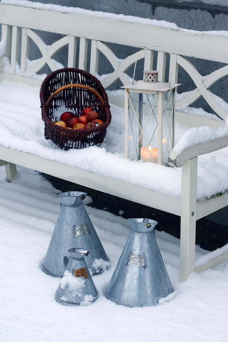 Korb mit Äpfeln und Laterne auf weißer Holzbank im Schnee