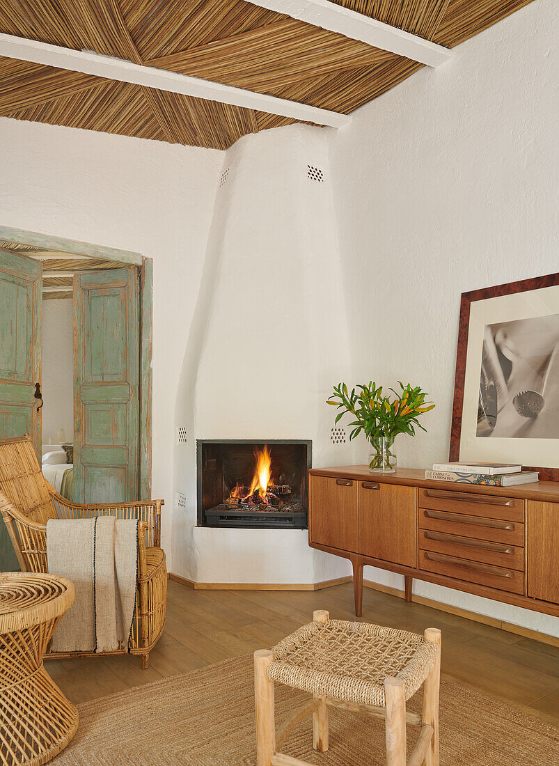 Sideboard und Rattansitzmöbel vor gemauertem Kamin im Wohnzimmer mit weißen Wänden