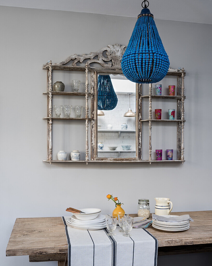 Rustikaler Holztisch mit Tischläufer und Geschirr, darüber blauer Kronleuchter und alter Bistroregal mit Spiegel