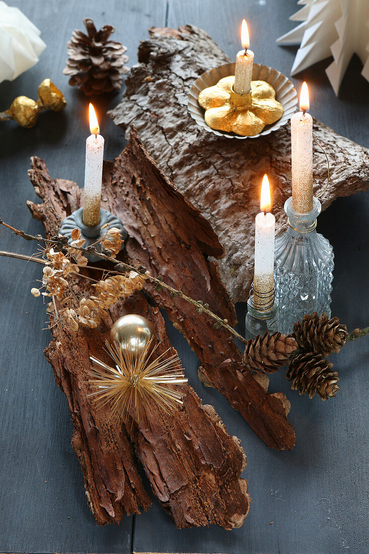 DIY-Adventskranz auf Baumrinde, dekoriert mit brennenden Kerzen auf grauem Holz und goldenem Weihnachtsbaumschmuck