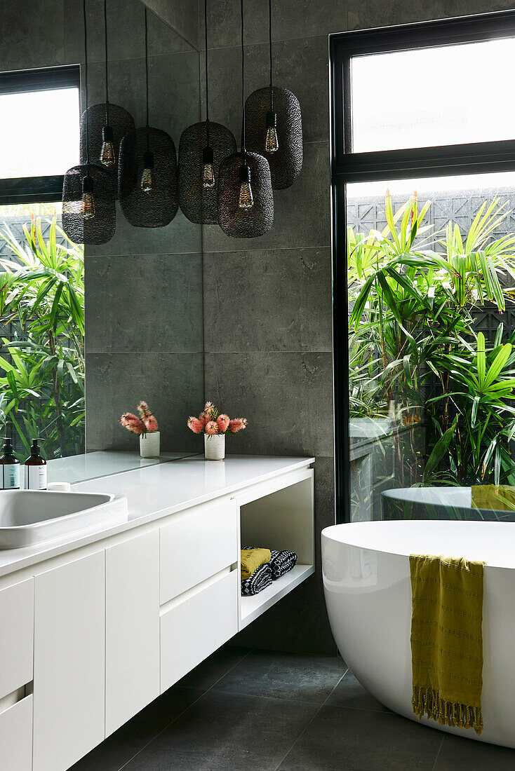 Free-standing bathtub in front of window, white washstand and black pendant lights in bathroom