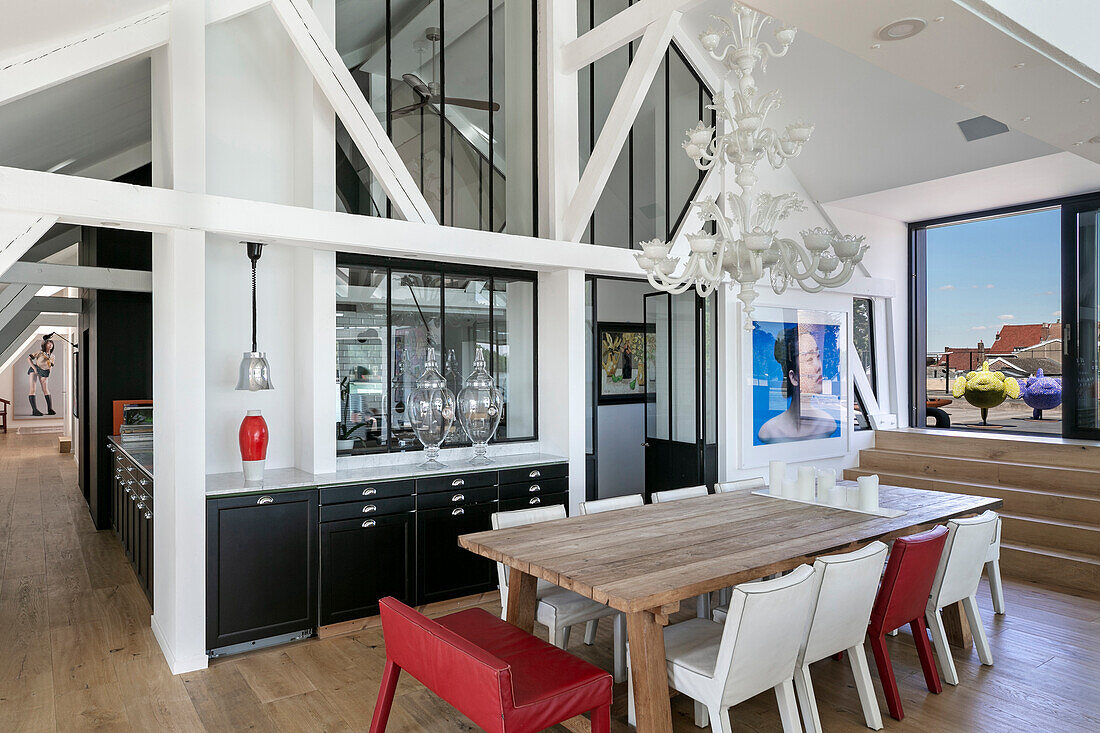 Wooden dining table with red and white chairs below chandelier in open-plan interior