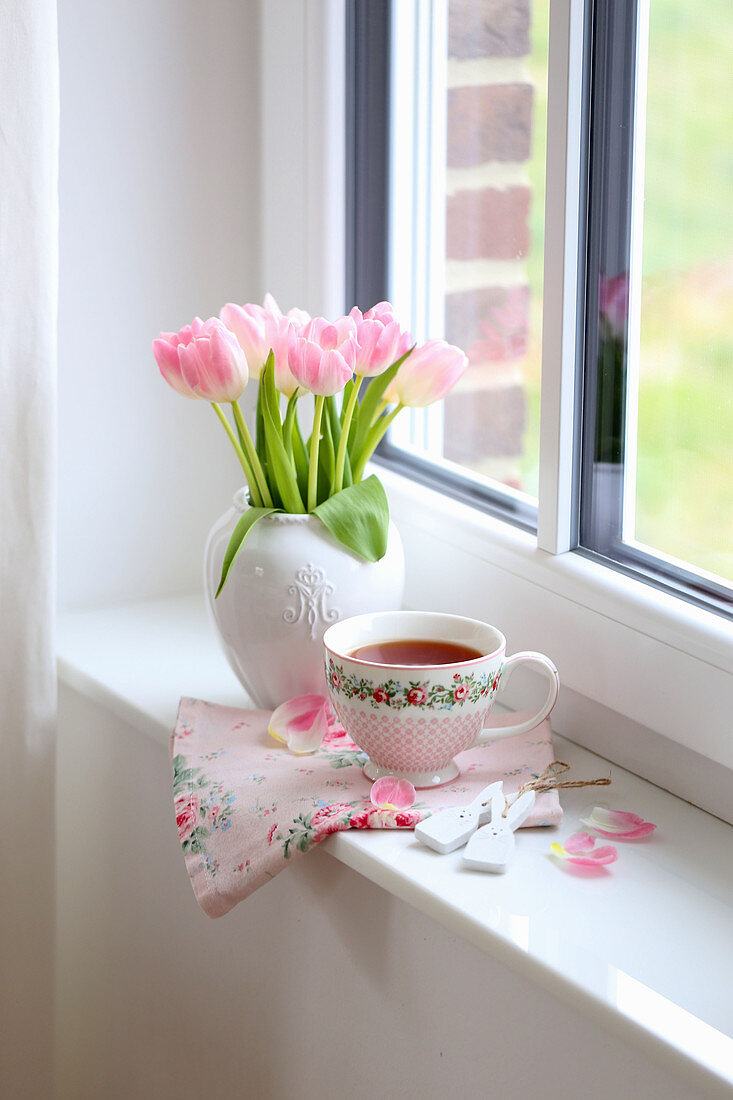 Vase with bouquet of tulips and teacup on windowsill