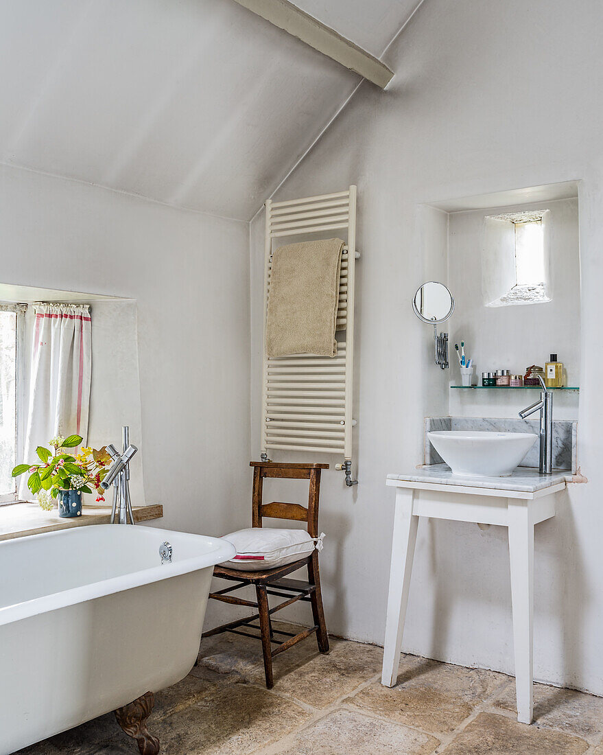 Bowl sink on white table, towel dryer, old chair, and freestanding bathtub in bathroom