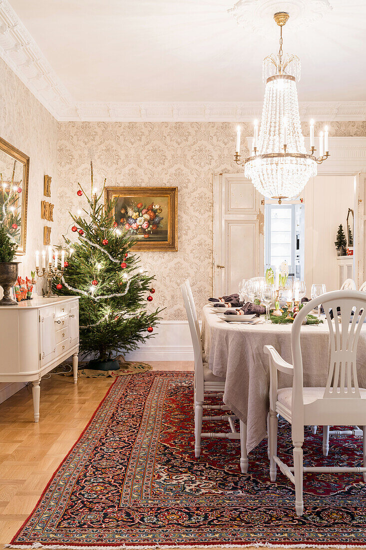 Festively set Christmas table in beige, chandelier above, Christmas tree in corner of room