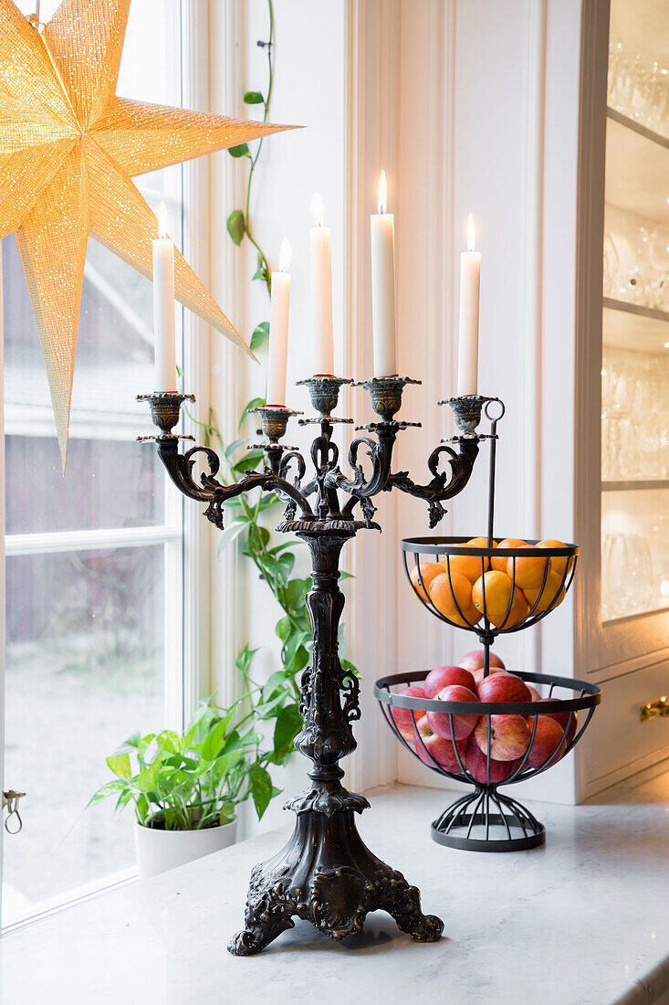 Kerzenhalter mit brenennden Kerzen und Obstkörbe auf Fensterbank, beleuchteter Papierstern am Fenster