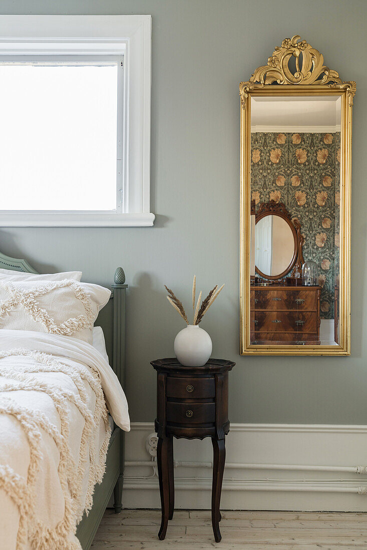 Gold-framed mirror and antique bedside table next to a bed in a bedroom with grey-green wall