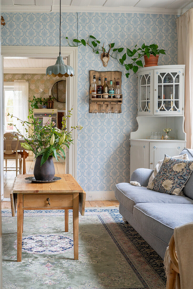 Wooden table with bouquet of flowers, sofa and corner cupboard in room with light blue wallpaper
