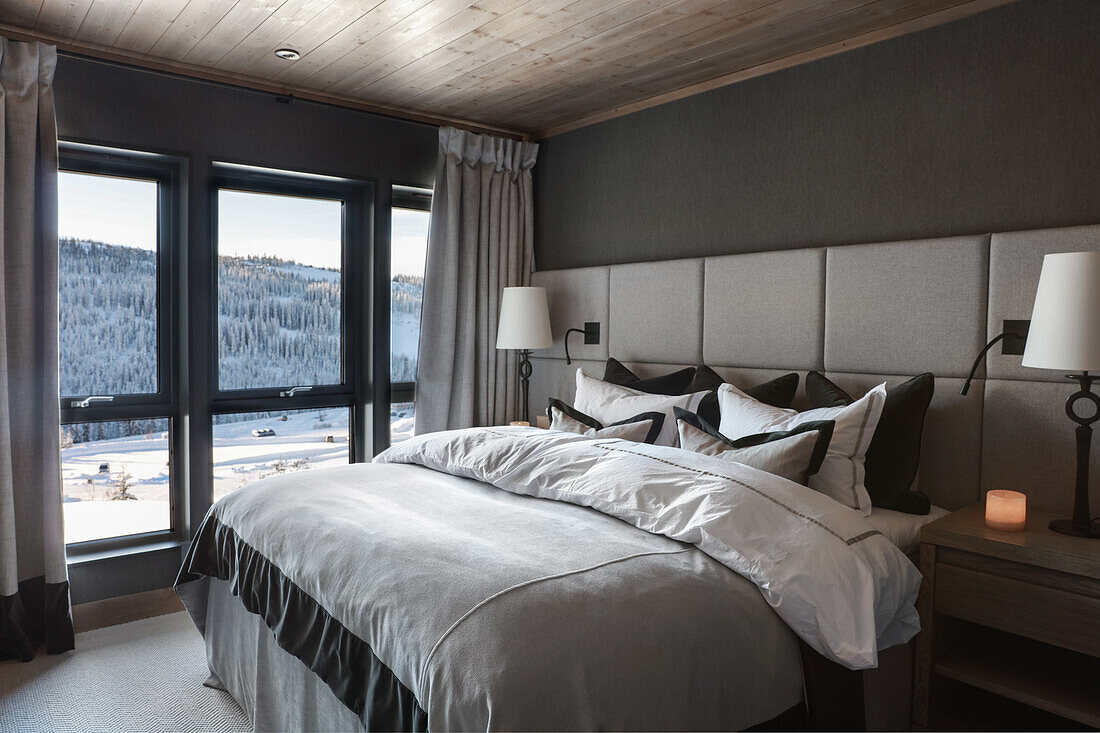 Doppelbett mit Betthaupt aus gepolstertem Paneel im Schlafzimmer, Fensterfront mit Blick auf schneebedeckte Landschaft