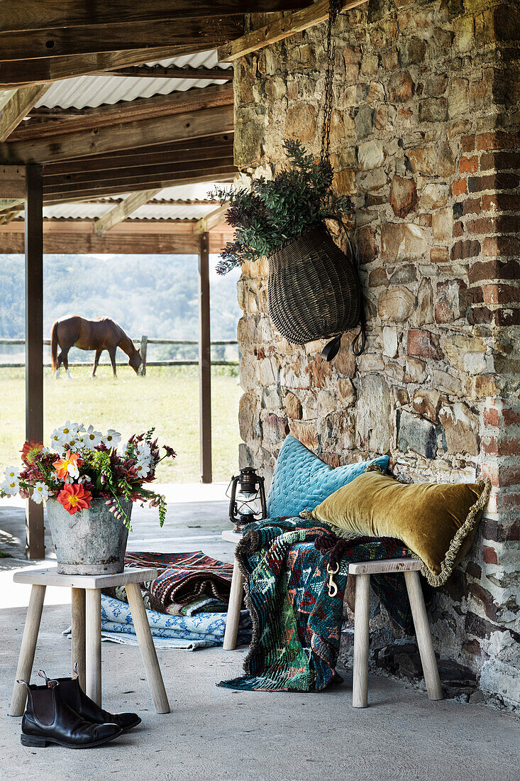 Überdachte Veranda mit Natursteinwand, Kissen und Teppich auf Holzbank, Eimer mit Blumenstrauß auf Hocker