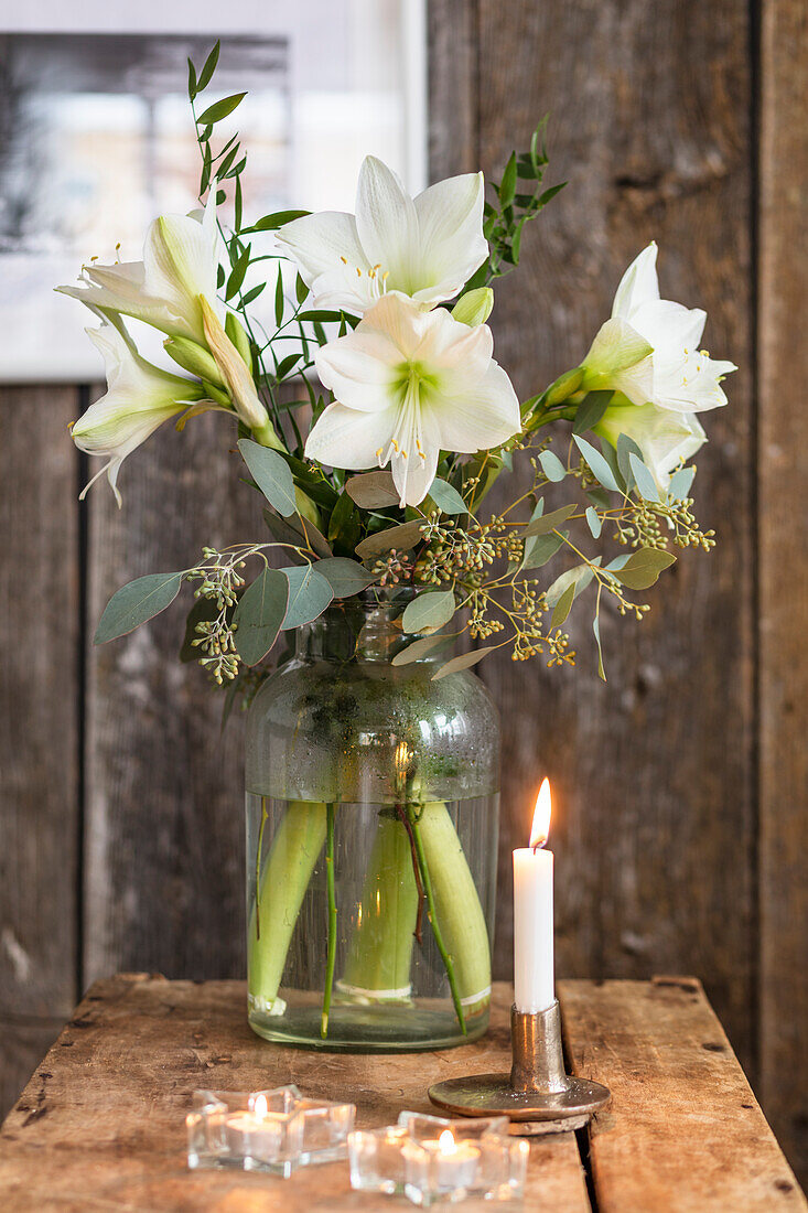 Weiße Lilien in Glasvase, Kerze und Teelichter auf Holzuntergrund und vor Holzwand