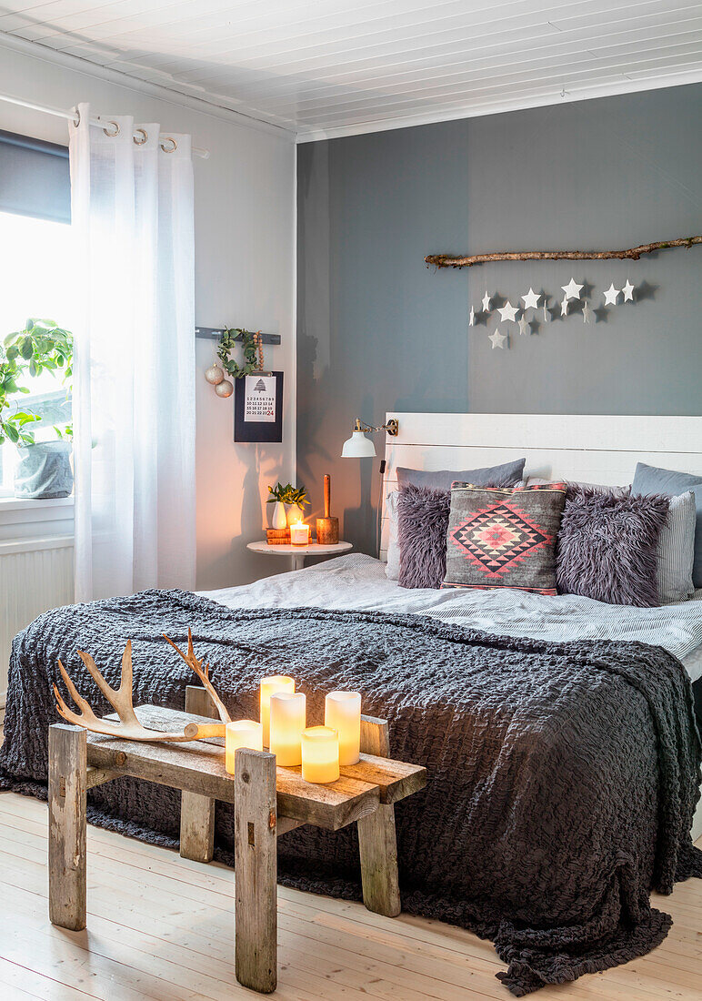 Bedroom with grey wall, candles on wooden bench, double bed with blankets and pillows and branch with star decoration on the wall