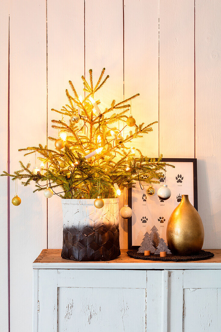 Christmas decoration with illuminated mini fir tree in a pot, golden vase and picture on chest of drawers