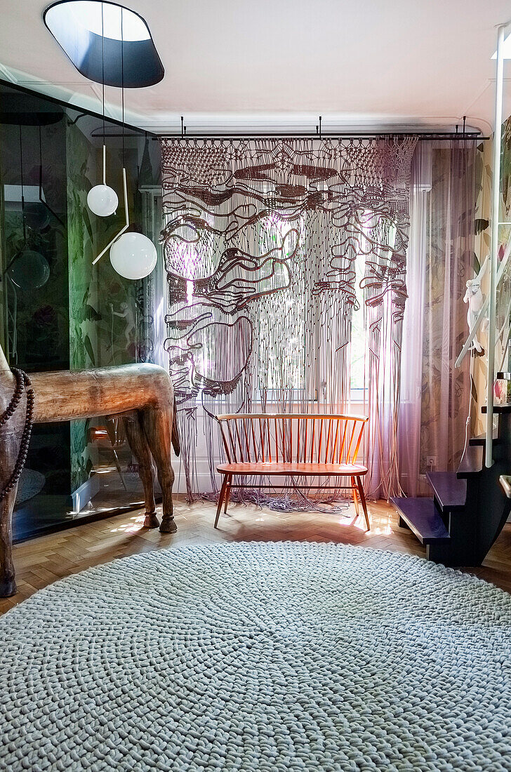 Wooden bench in front of window with patterned curtains and round knitted rug in eclectic living room