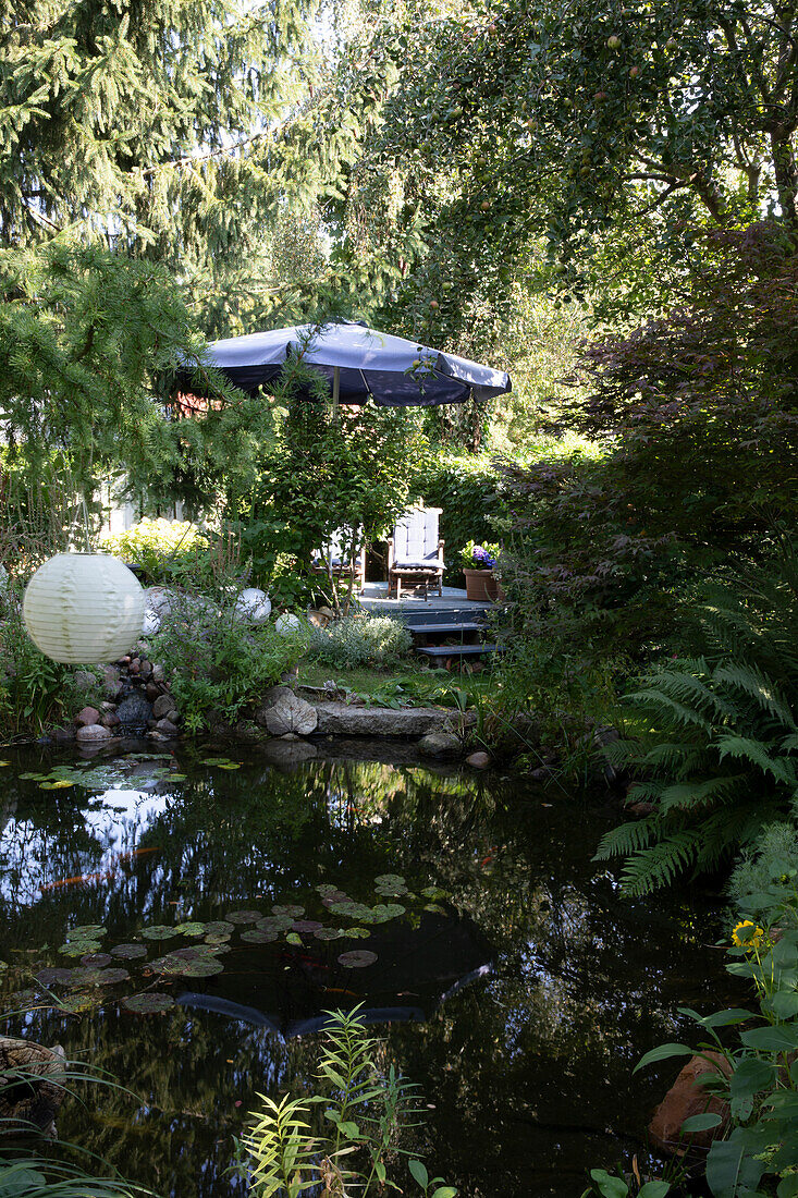 Laterne über Teich im üppig bewachsenen Garten, im Hintergrund Terrassenplatz mit Sonnenschirm