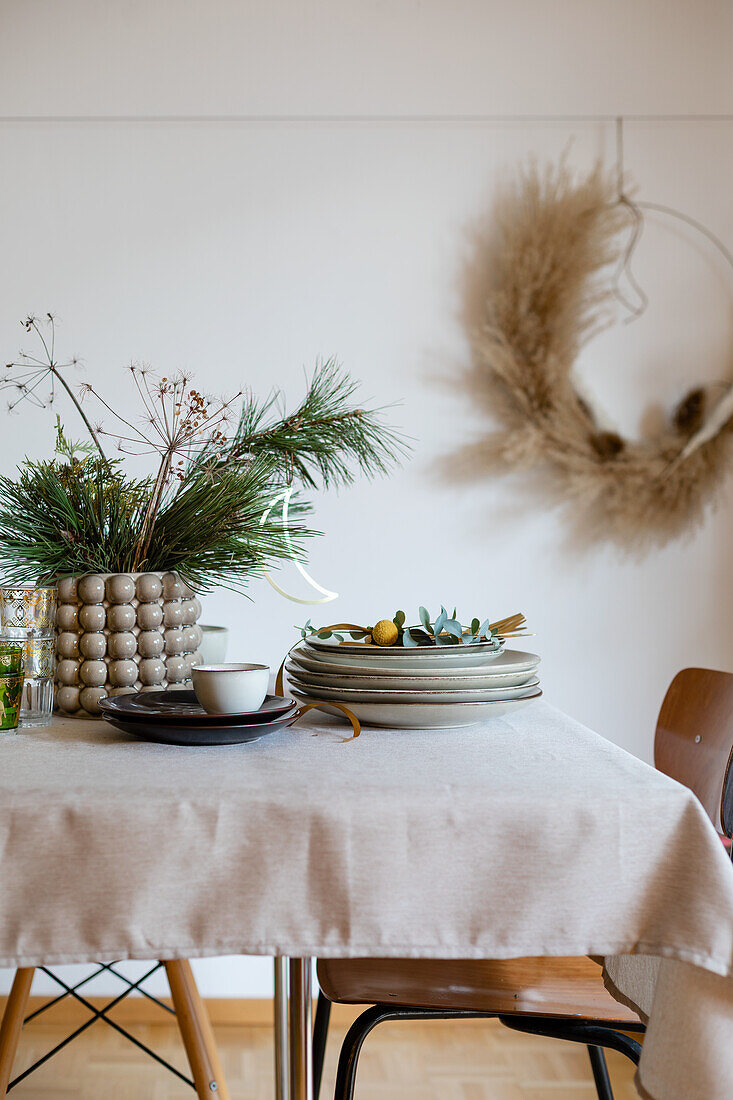 Materials for setting a Christmas dining table: vase with pine branches, stack of plates and golden cutlery, floral hoop on the wall