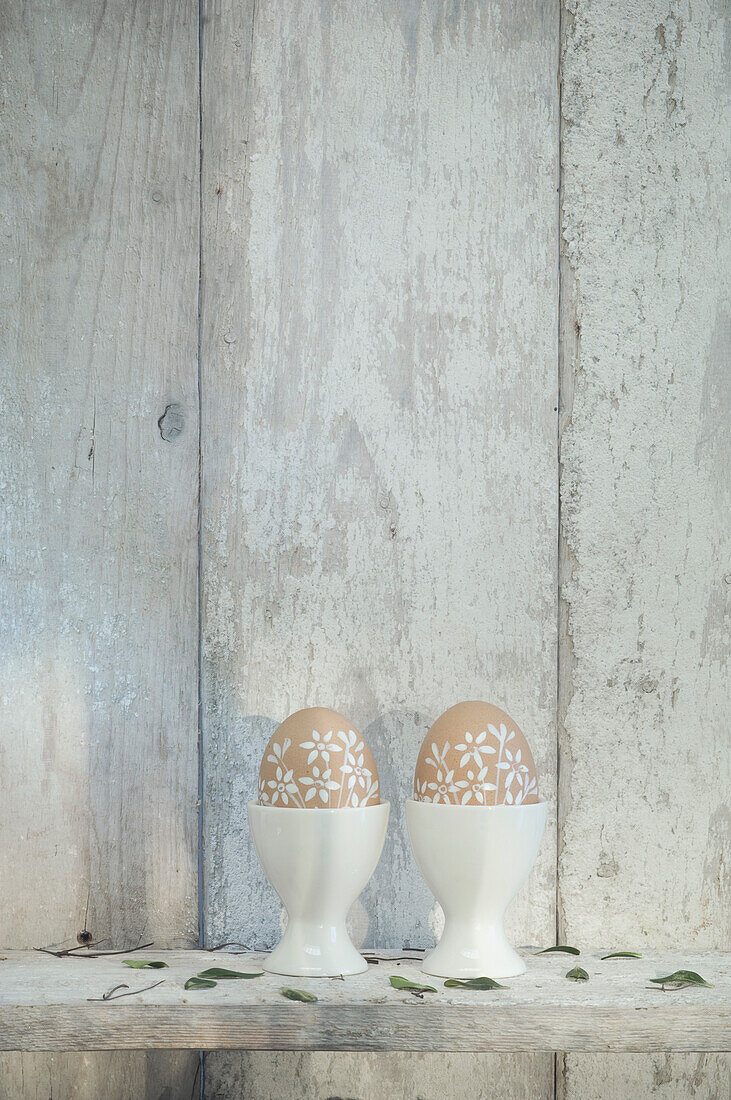 Two painted Easter eggs in egg cups on shelf in front of wooden wall
