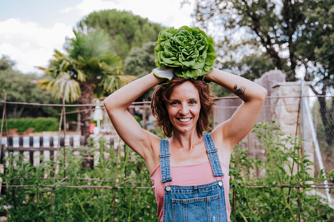 Smiling woman holding lettuce on head while standing in vegetable garden