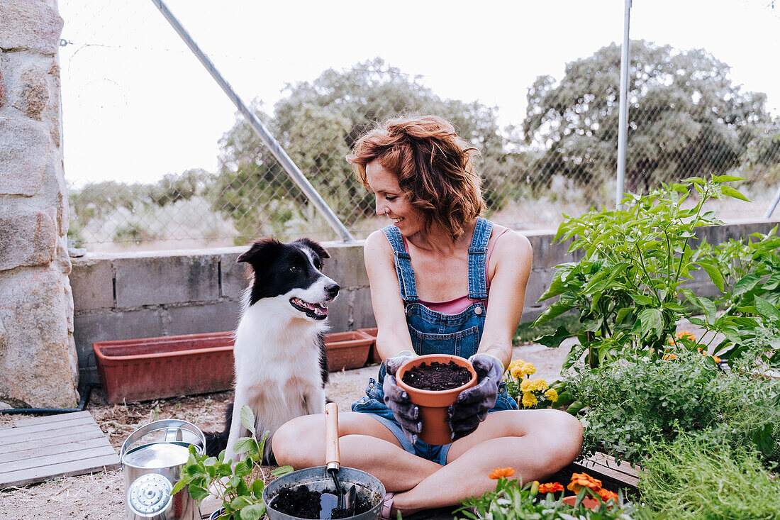 Lächelnde Frau mit Blumentopf und Blick auf Border Collie, während sie im Gemüsegarten sitzt