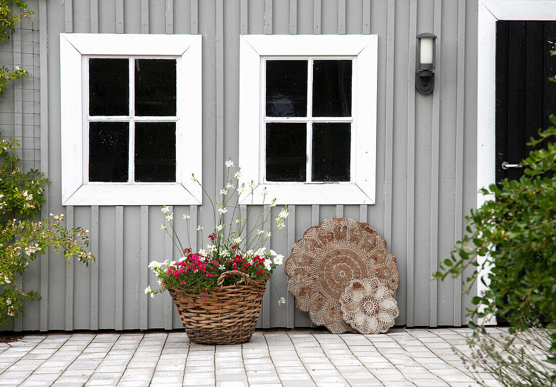 Graues Holzhäuschen mit weißen Fensterrahmen, davor Blumenkorb und dekorative Betonplatten