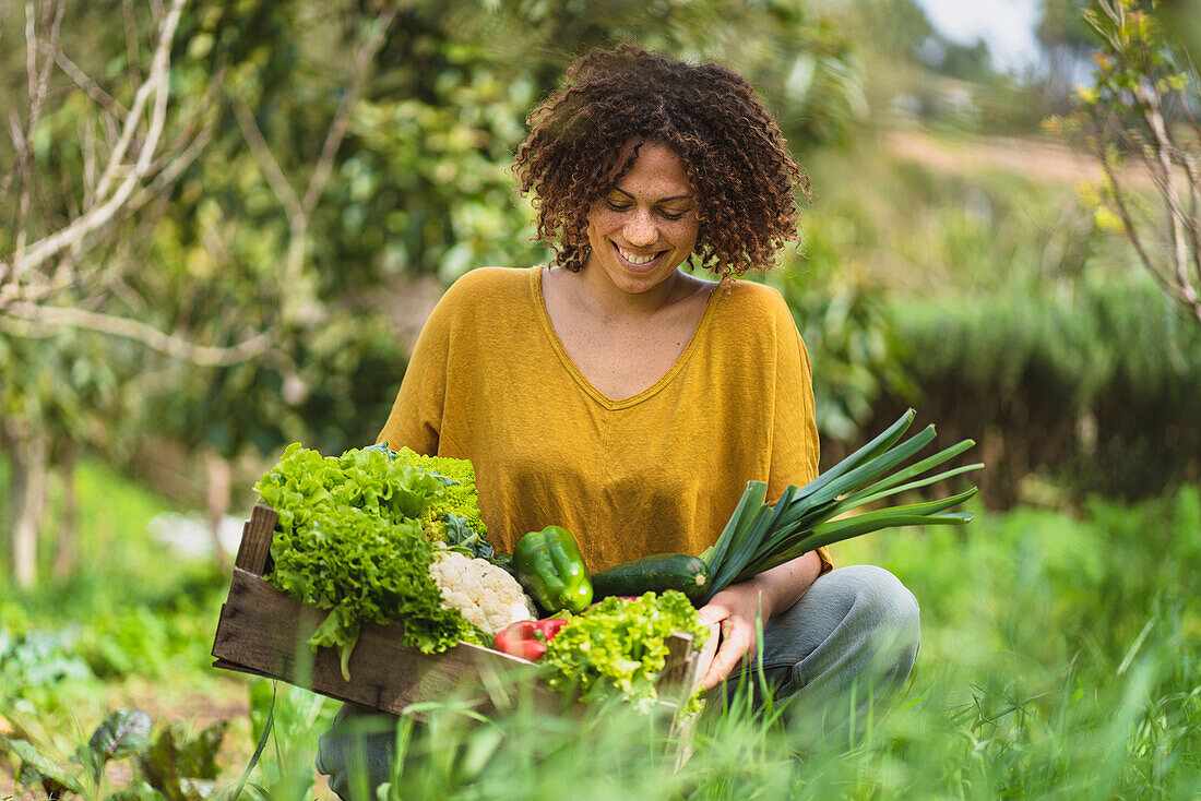 Lächelnde Frau, die Gemüse in einer Kiste betrachtet, während sie im Garten hockt