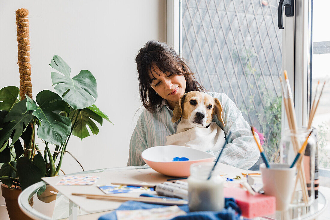 Attractive female painter caressing cute Beagle dog while sitting at table with painted bowl and various paintbrushes