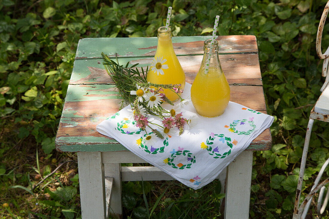 Zwei Flaschen mit Orangengetränk, Wildblumen und Tischdecke auf Holzhocker