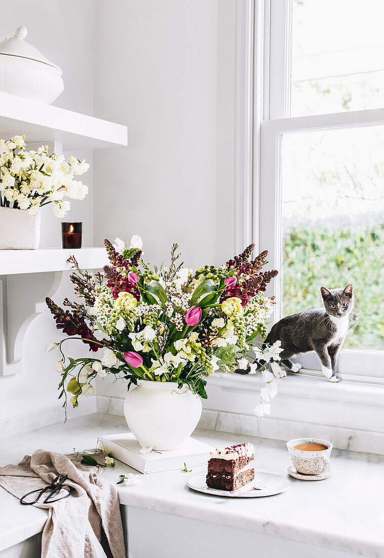 Üppiger Blumenstrauß, ein Stück Kuchen mit Kaffee vor Fenster, Katze auf der Fensterbank