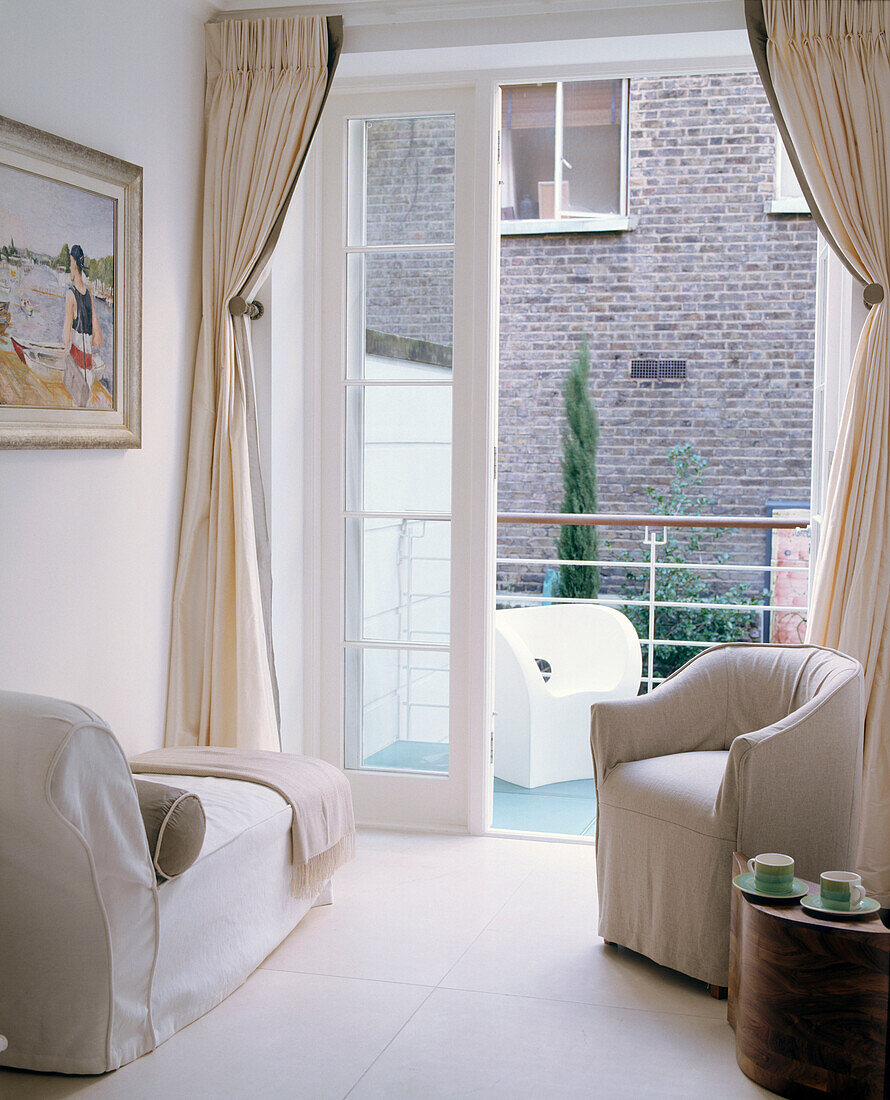 Ein traditionelles, in neutralen Farben gehaltenes Wohnzimmer mit gepolstertem Chaiselongue-Sessel und französischen Fenstern, die auf einen Balkon führen