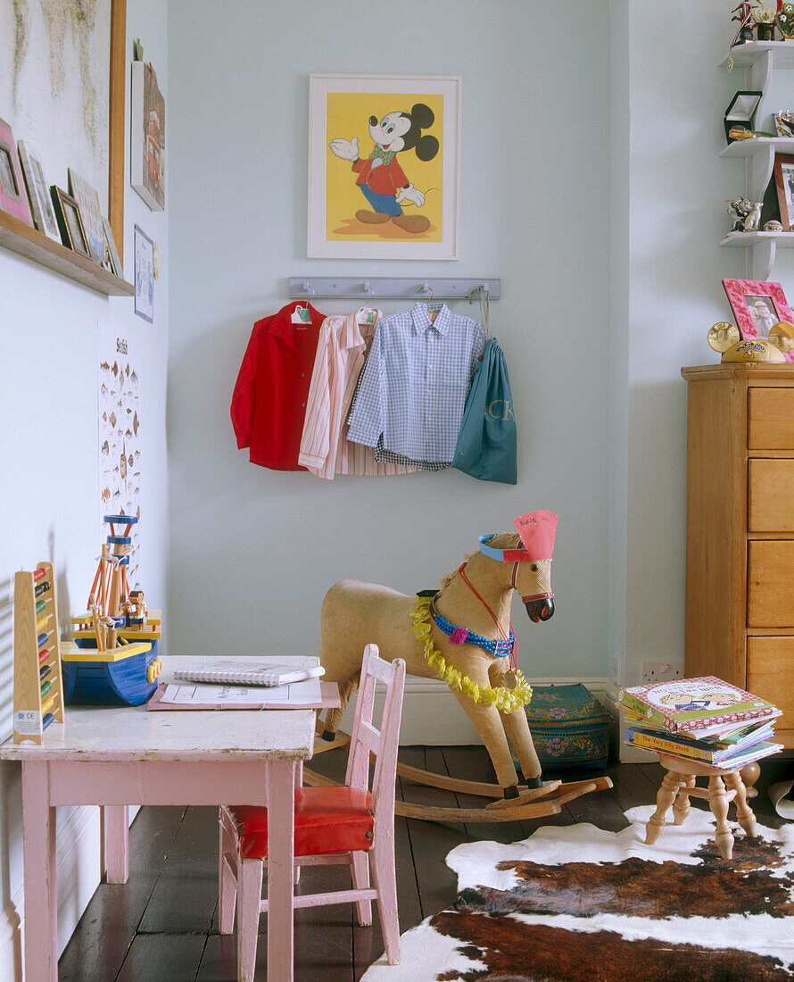 A traditional child's bedroom with a framed cartoon above a row of coat pegs behind a rocking horse and a pink table and chair