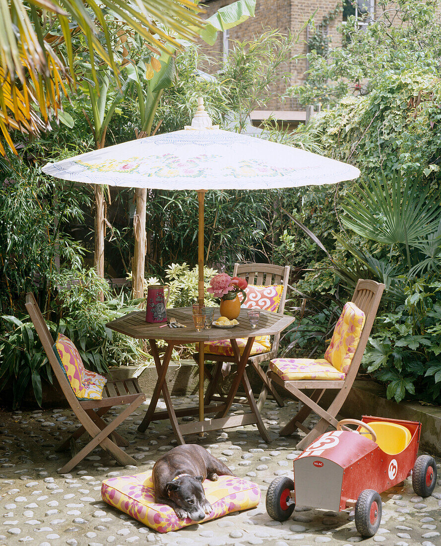 An enclosed garden with a dog lying on a cushion next to a toy car wooden table and chairs with a parasol surrounded by plants and trees
