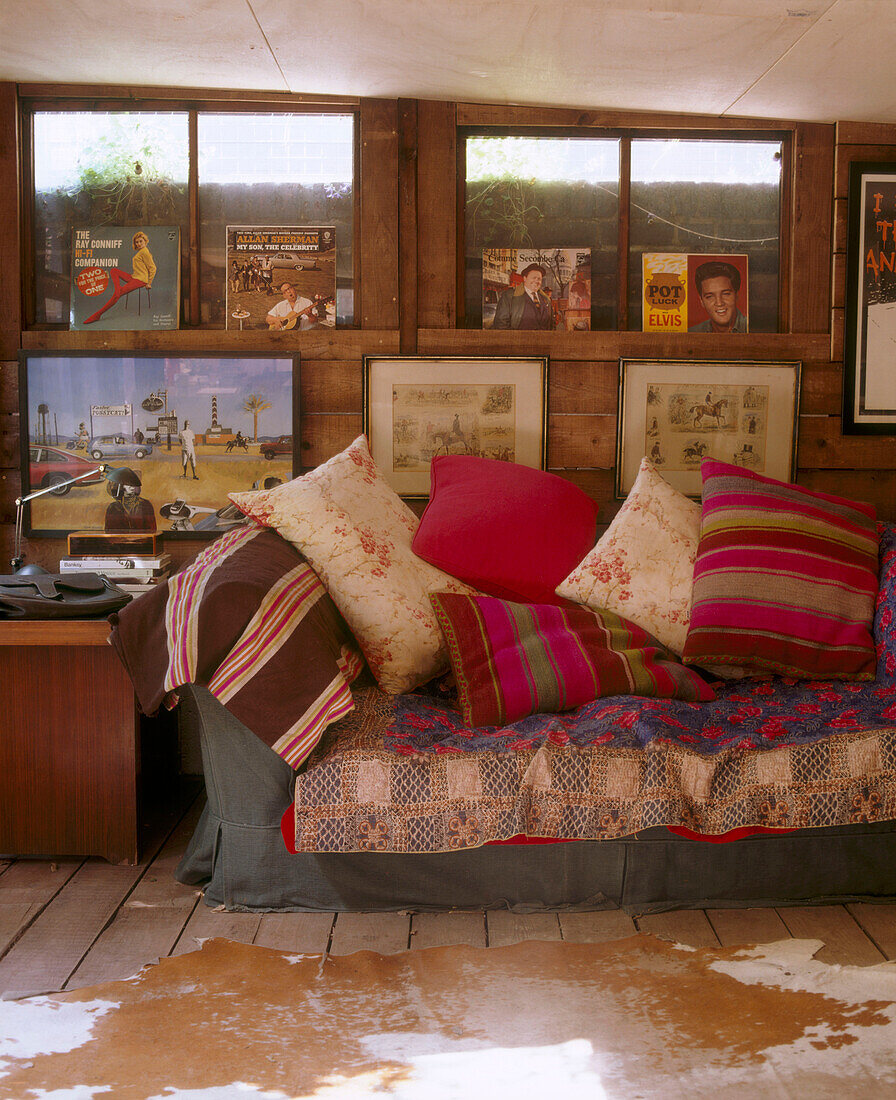 Detail einer Wand mit gerahmten Bildern und Schallplatten über einem traditionellen Sofa mit einer Vielzahl von Kissen vor einem Teppich