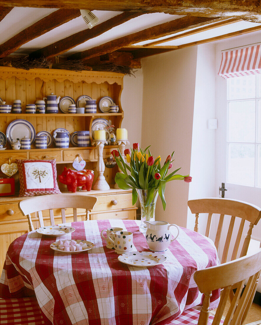 Ein Esszimmer im Landhausstil mit rundem Küchentisch mit karierter Tischdecke und Holzstühlen vor einer großen Kommode aus Kiefernholz, auf der eine Vielzahl von Geschirr steht