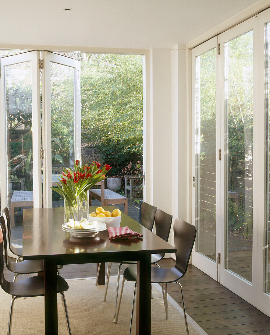 Esszimmer im Wintergarten mit Glaswänden darin moderner Esstisch umgeben von Stühlen und gedeckt mit Speisen und Blumen