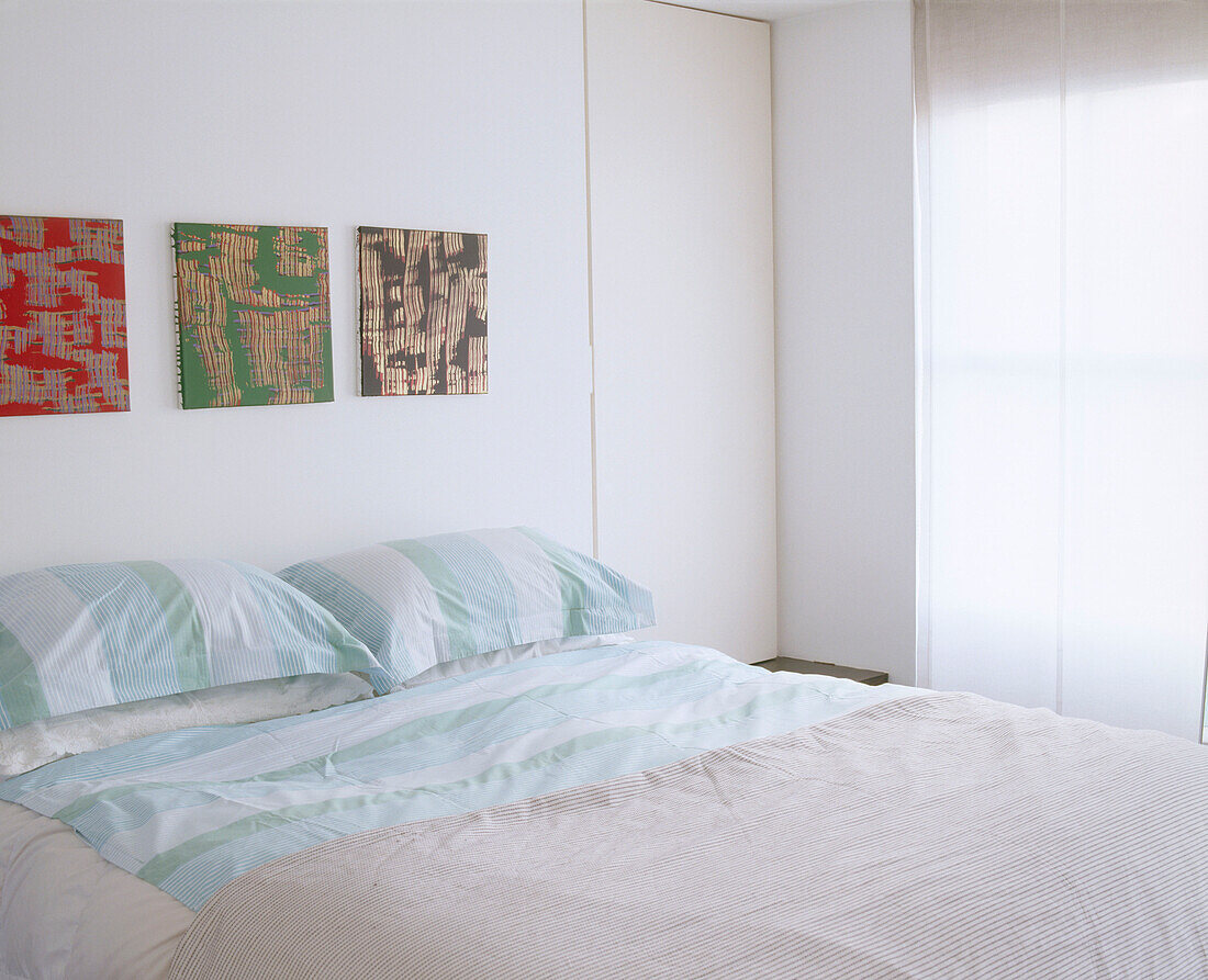 A modern minimalist bedroom with three canvas paintings mounted above a the head of a bed and next to a large window with a blind