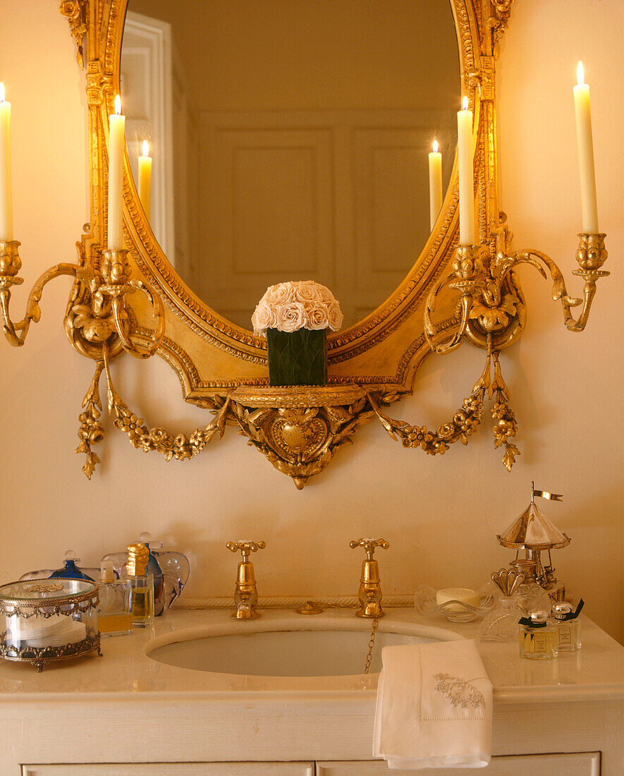 Ein Detail eines modernen Badezimmers mit einem Waschbecken in einem Schrank und einem verschnörkelten Goldspiegel mit Kandelabern