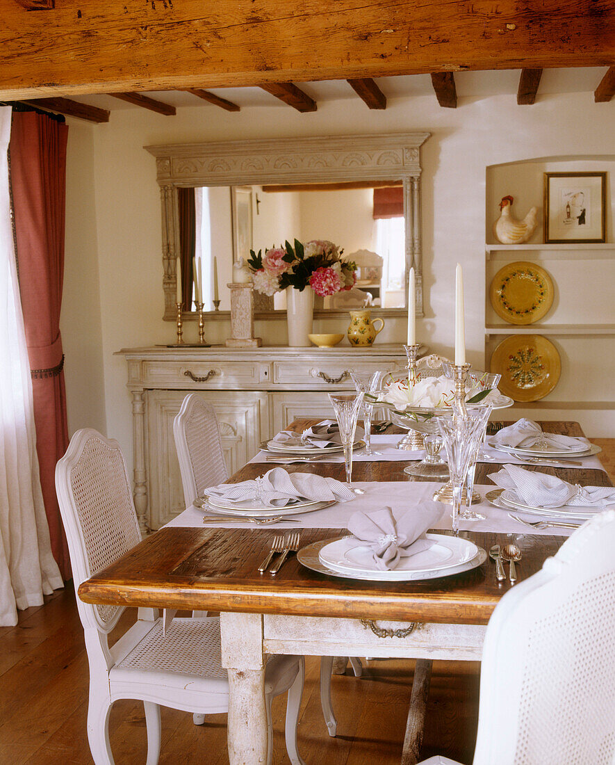 A country style dining room with a place settings on a wooden table surrounded by white painted chairs below large exposed wooden beams