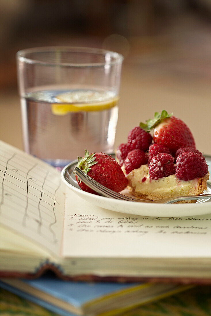 Teller mit sommerlicher Obsttorte auf aufgeschlagenem Buch dahinter ein Glas Wasser, Richmond upon Thames, London, UK