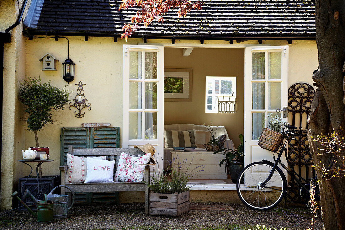 Sitzbank mit bestickten Kissen und ein Fahrrad neben geöffneten Flügeltüren an einem Haus in West Sussex, England, UK