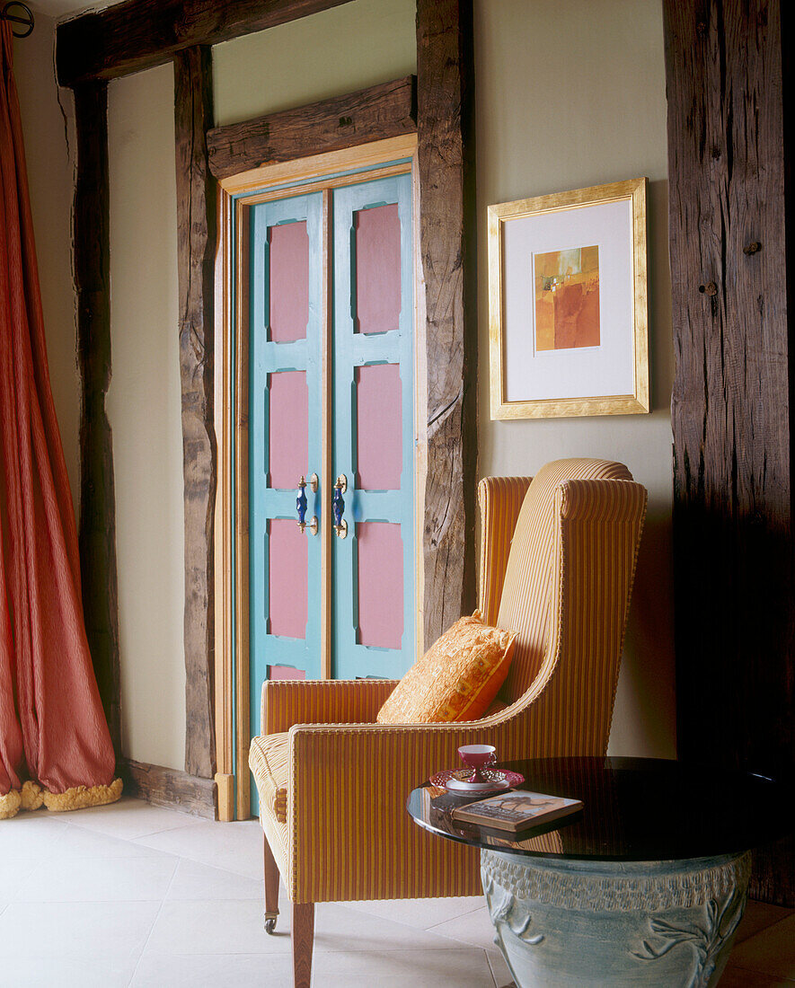 Traditioneller Sessel mit orangefarbener Stoffpolsterung neben einer kleinen Doppeltür mit blauem und rosa Holzrahmen