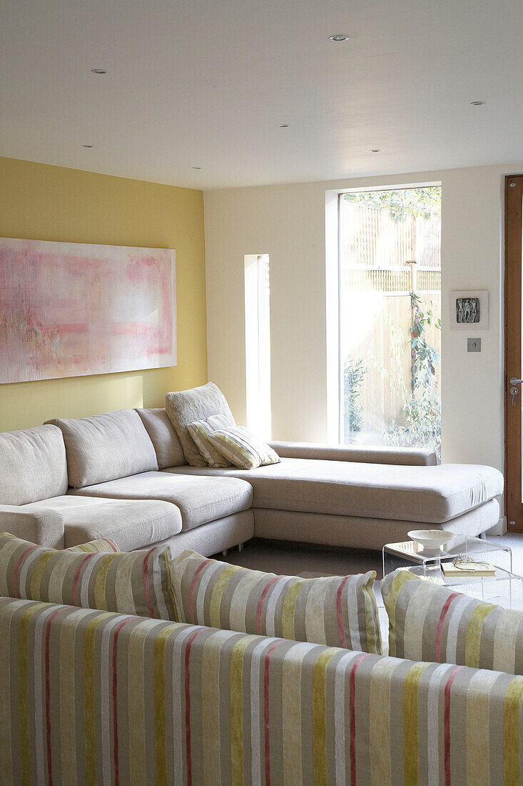 Ein modernes Wohnzimmer mit einem großen L-förmigen Sofa unter einem rosa abstrakten Gemälde neben einem großen Fenster