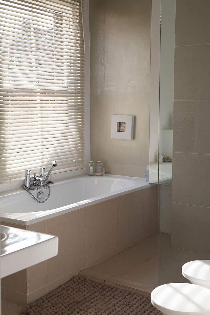 Ein modernes Badezimmer mit einem Fenster mit Jalousien mit Blick auf die von beigen Fliesen umgebene Badewanne
