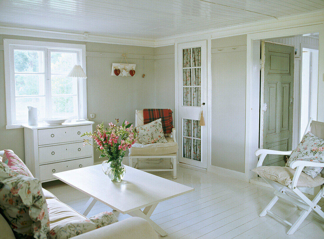 Wohnzimmer im Landhausstil in neutralen Farben, mit gestrichenen Dielen, Stühlen, Kommode, Polsersofa, geblümten Stoffen und Blumenarrangement