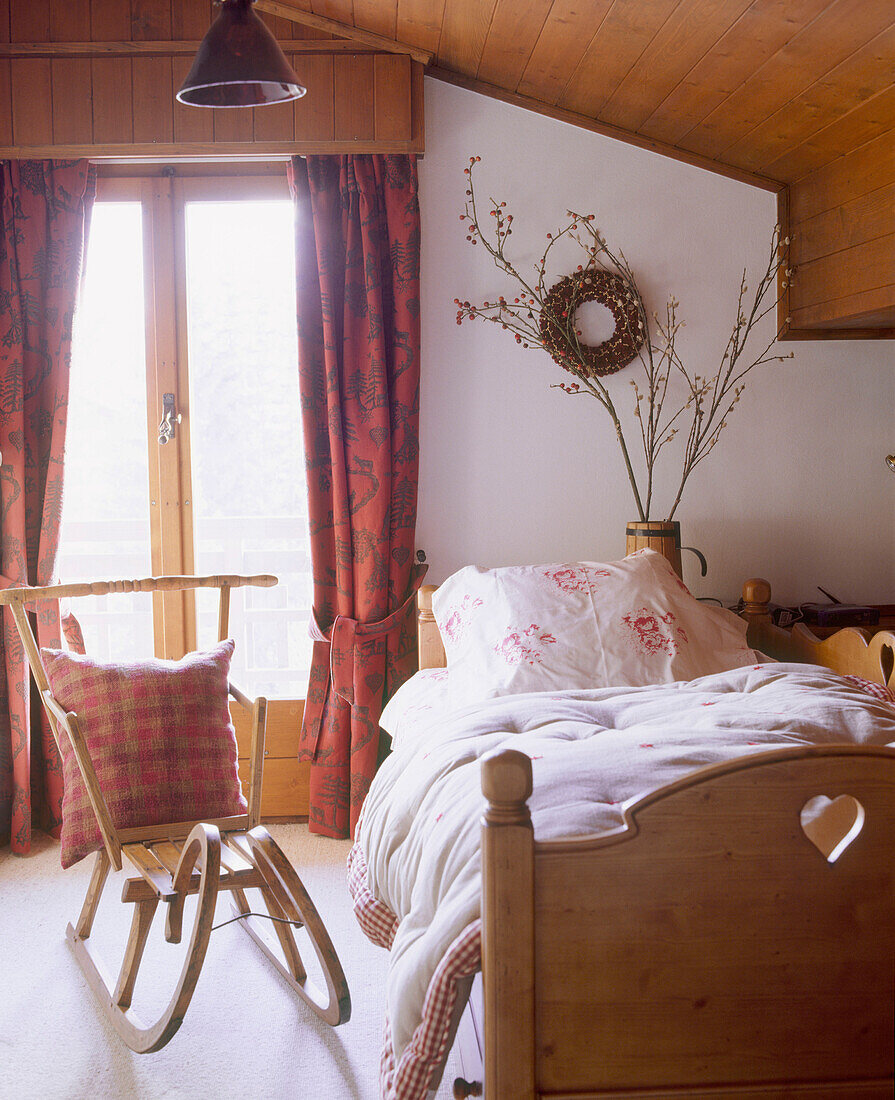 Ein Schlafzimmer im Landhausstil mit holzgetäfeltem Einzelbett und rot gemusterten Vorhängen sowie einem altmodischen Schlitten als Sitzgelegenheit