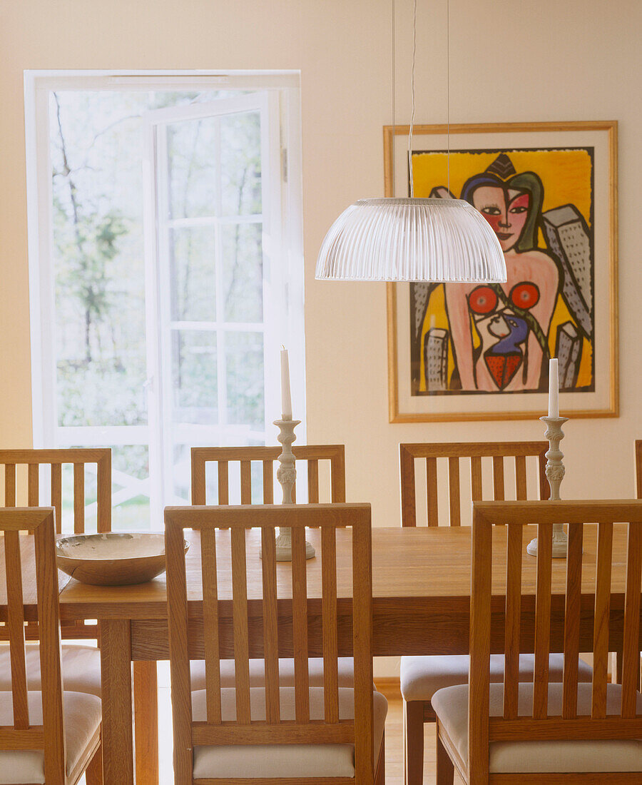 Glaslampenschirm über Esstisch und Stühlen aus Holz dahinter gerahmtes Wandgemälde und geöffnete Terrassentüren