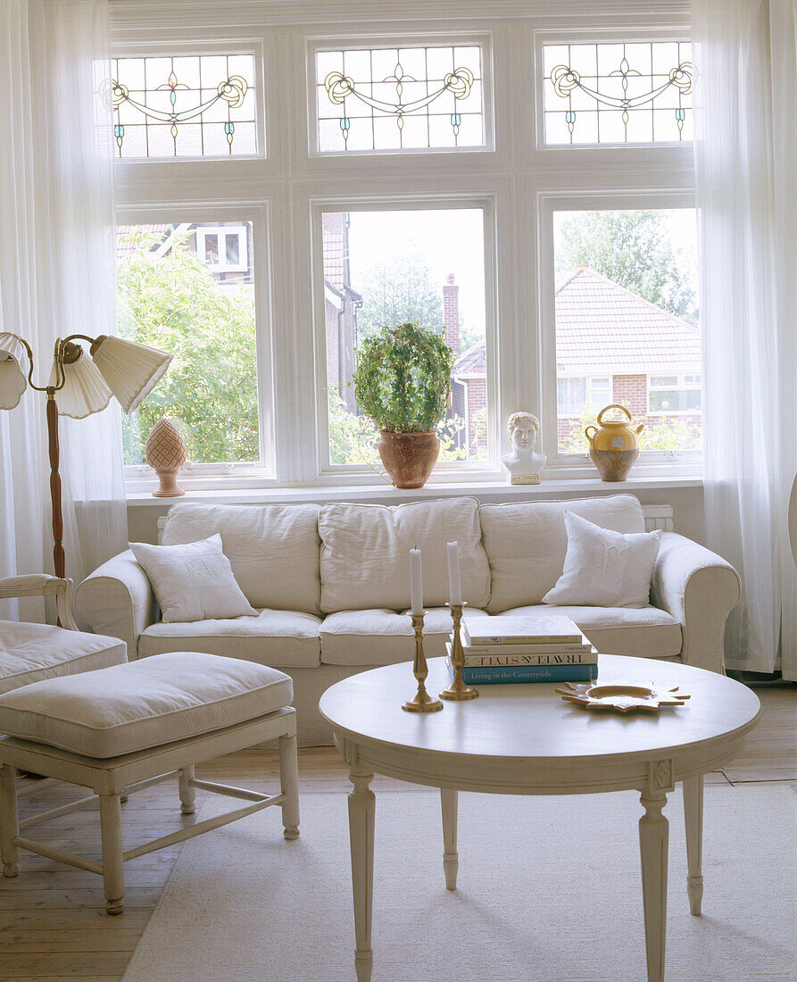 Ein traditionelles Wohnzimmer in Weiß mit Sofa, rundem Tisch und Fußbank vor großem Fenster
