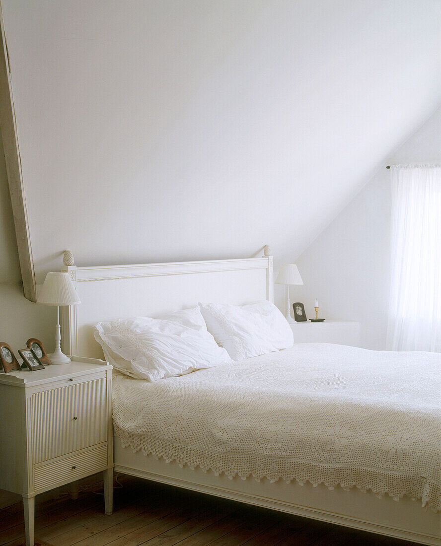 A traditional bedroom featuring a sloped ceiling with white walls and bed linen next to a small bed side table