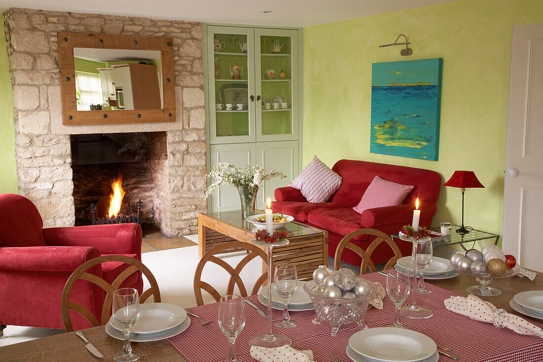 Ein offenes, modernes, grünes Wohnzimmer im Landhausstil mit einem Steinkamin, in dem ein roter Polstersessel brennt, und einem Sofatisch, an dem Kerzen für das Abendessen brennen