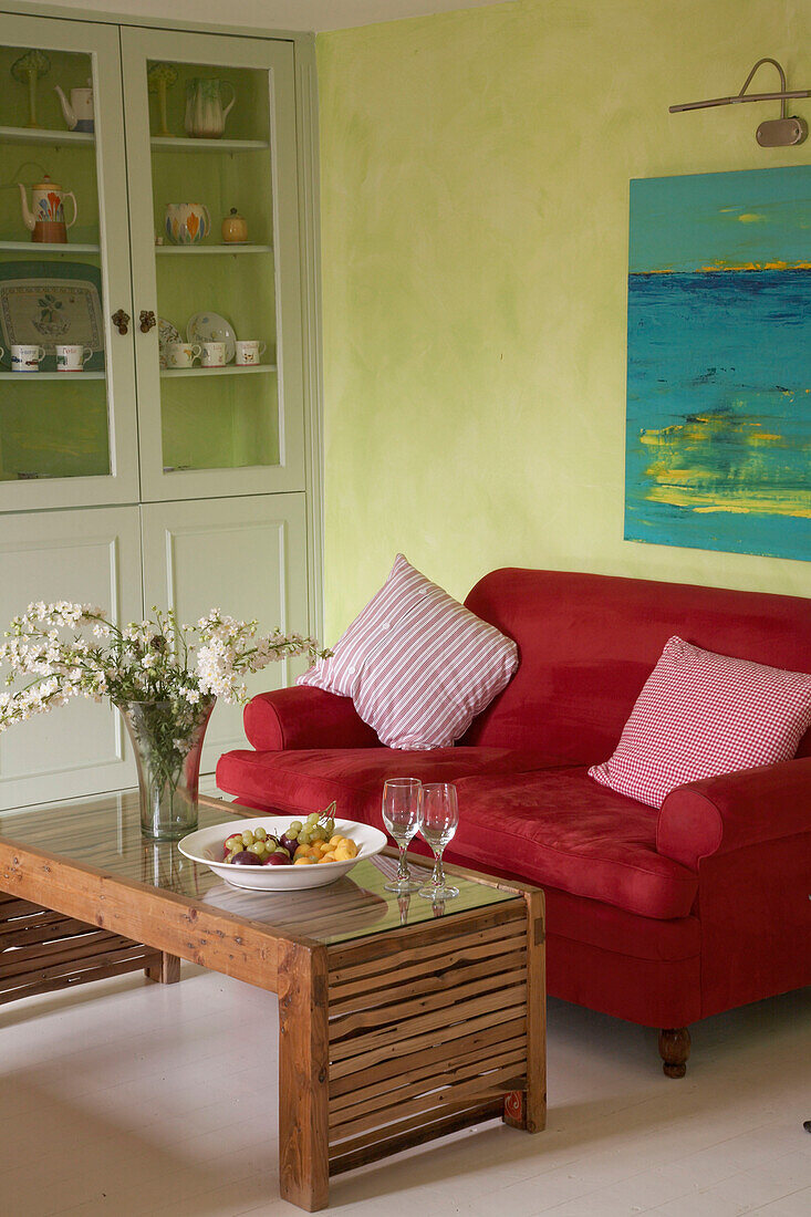 Ein offenes, modernes, grünes Wohnzimmer im Landhausstil mit einem rot gepolsterten Sofa, einem Couchtisch aus Holz, Gläsern, Kissen und Blumenarrangements in einem Vitrinenschrank